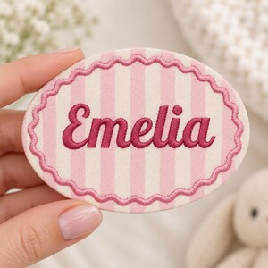 May include: An oval-shaped fabric patch featuring a light pink and white vertical striped pattern. The name Emelia is embroidered in dark pink cursive script in the centre, surrounded by a scalloped dark pink embroidered border edge.