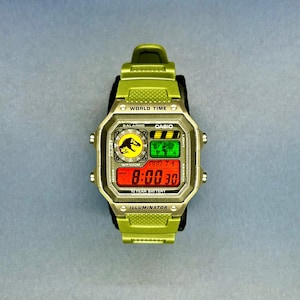 Op de afbeelding: Een groene Casio digitale horloge met een zwarte band. De wijzerplaat toont de tijd, datum en diverse functies. Het horloge heeft het Jurassic Park-logo en de tekst "WORLD TIME", "BALARMS", "CASIO", "WR100M", "10 YEAR BATTERY" en "ILLUMINATOR".