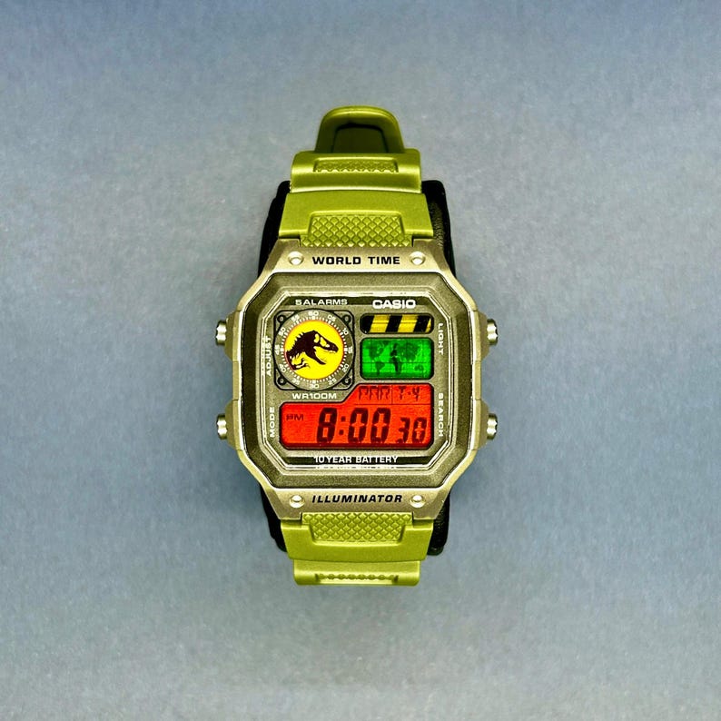May include: A green Casio digital watch with a black band. The watch face displays the time, date, and various functions. The watch features the Jurassic Park logo and the text "WORLD TIME", "BALARMS", "CASIO", "WR100M", "10 YEAR BATTERY", and "ILLUMINATOR".