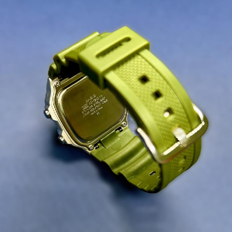 May include: A green digital watch with a silver-tone case and a matching green strap. The watch has a rectangular face and a buckle closure. The back of the watch has text and is made of stainless steel.