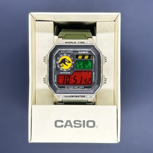 May include: A Casio digital watch with a green strap and a silver-colored case, displayed in a white box. The watch face features a dinosaur logo, world map, and a red digital display showing the time. The watch has the text "WR100M" and "10 YEAR BATTERY".