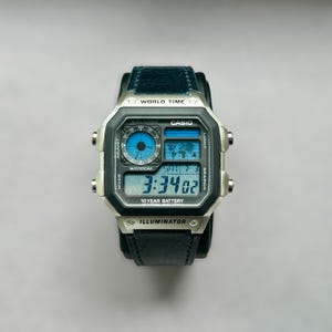 Op de afbeelding: Een zilveren Casio digitale horloge met een zwarte leren band. De wijzerplaat toont de tijd, datum en wereldtijdfuncties. Het horloge heeft de tekst "CASIO", "WR100M", "ILLUMINATOR" en "10 YEAR BATTERY".