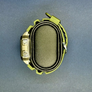 May include: A digital watch with a silver face and olive green strap. The watch has two buttons on the left side and a buckle closure. The watch is displayed on a black oval base.