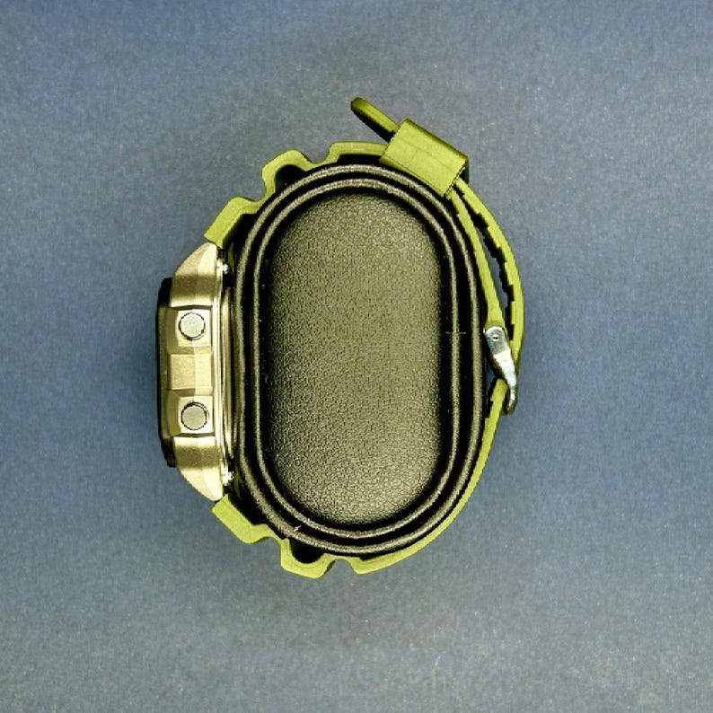 May include: A digital watch with a silver face and olive green strap. The watch has two buttons on the left side and a buckle closure. The watch is displayed on a black oval base.