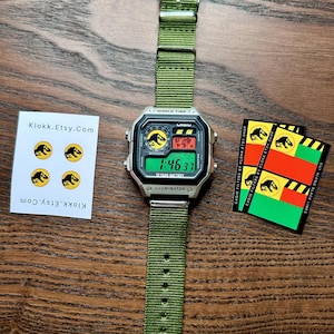 May include: A silver Casio digital watch with a green nylon strap, featuring a Jurassic Park logo on the face. The watch displays the time 1:46:37. Also shown are stickers with the Jurassic Park logo and the text "Klokk.Etsy.Com".