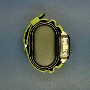 May include: An olive green digital watch with a black face and silver-colored buttons. The watch has a matching green strap and is displayed on a black oval watch holder. The watch is a modern accessory.