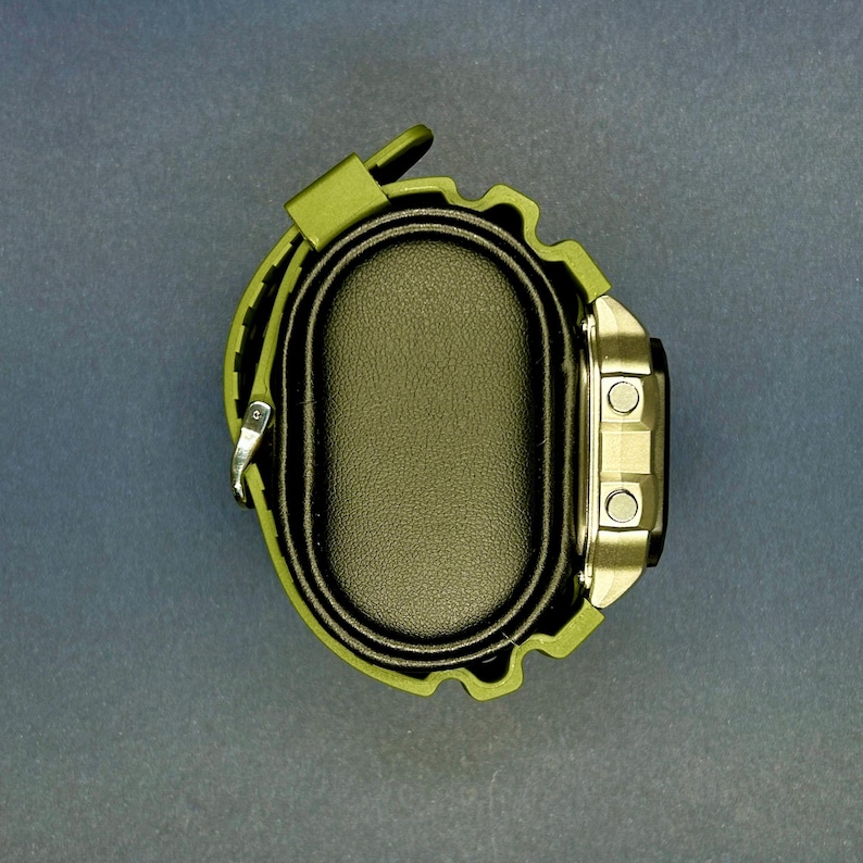May include: An olive green digital watch with a black face and silver-colored buttons. The watch has a matching green strap and is displayed on a black oval watch holder. The watch is a modern accessory.
