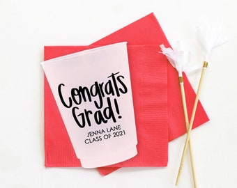 May include: A pink plastic cup with the text "Congrats Grad!" and "Jenna Lane Class of 2021" printed on it. The cup is sitting on top of two red napkins.