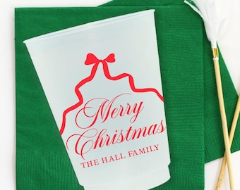May include: A frosted plastic cup with red text reading "Merry Christmas" and "THE HALL FAMILY" is displayed on a green napkin. Two decorative sticks with white feather accents are also present. The items are arranged on a white surface.