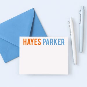 May include: A white rectangular note card featuring the name HAYES PARKER printed in orange and blue, accompanied by a light blue envelope and two white pens. The card is blank below the printed name, providing space for personal written messages.