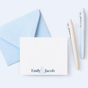 May include: A white rectangular note card featuring the printed text Emily & Jacob in dark blue ink, accompanied by a light blue envelope and two ballpoint pens, one in a light beige tone and the other in a light blue shade.
