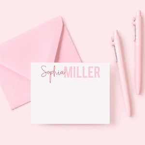 May include: A white rectangular note card featuring the name Sophia Miller printed in a combination of script and block lettering in a pink hue, accompanied by a light pink envelope and two matching light pink pens positioned to the right.