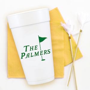 May include: White cup with green text and a golf flag graphic, resting on a yellow napkin. The text reads "The Palmers". Two decorative feather sticks are also present.