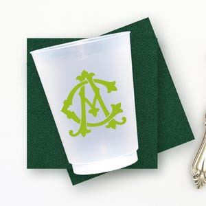 May include: A clear plastic cup with a green monogram, resting on a dark green square napkin. A silver bottle opener with an ornate handle is positioned to the right. The items are on a white surface.