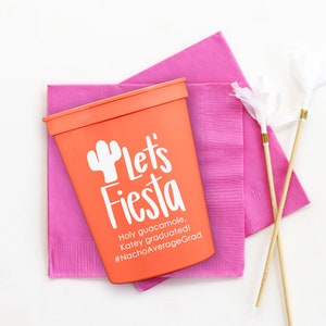 May include: Orange plastic cup with the text "Let's Fiesta" and a cactus graphic. The text below reads "Holy guacamole. Katey graduated! #NachoAverageGrad". The cup is sitting on top of two pink napkins and two wooden skewers with white feathers.