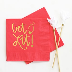 May include: Red paper napkins with gold lettering that reads "Get Lit!" and a lightbulb graphic. Two white and gold decorative sticks are also in the image. The napkins are square and have a textured surface.
