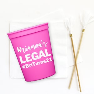 May include: A pink plastic cup with the text "Brianna's LEGAL #BritTurns21" printed on it. The cup is sitting on a white napkin with two wooden skewers with white tassels.