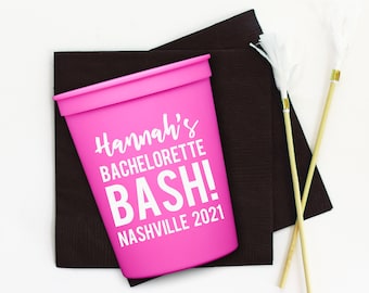 May include: A bright pink plastic cup with the text "Hannah's BACHELORETTE BASH! NASHVILLE 2021" in white. The cup sits on a dark brown napkin, with two decorative feather wands with gold accents.