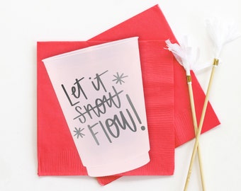 Puede incluir: Un vaso de plástico transparente con el texto "Let it snow FLOW!" en negro, sobre una servilleta roja. También hay dos agitadores de cóctel decorativos con detalles de papel blanco y bandas doradas. El fondo es blanco.