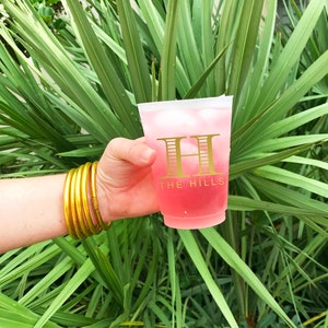 May include: A pink plastic cup with ice and a pink beverage. The cup has a gold design with the text "THE HILLS" and a stylized letter "H".