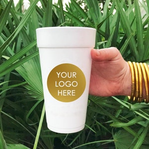 May include: White foam cup with a gold circle that says "YOUR LOGO HERE" on it. The cup is being held by a hand with gold bangles.