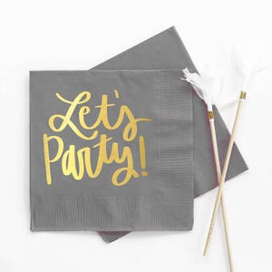 Op de afbeelding: Grijze papieren servetten met gouden letters die "Let's Party!" zeggen.