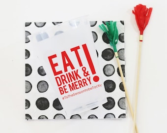 May include: A clear plastic bag with the text "EAT! DRINK & BE MERRY" in red, placed on a white napkin with black polka dots. Two decorative sticks with red and green accents are also present.
