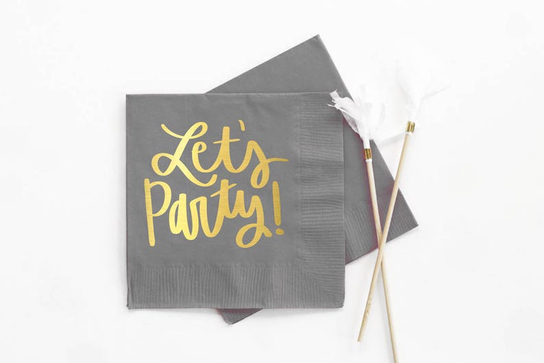 Op de afbeelding: Grijze papieren servetten met gouden letters die "Let's Party!" zeggen.