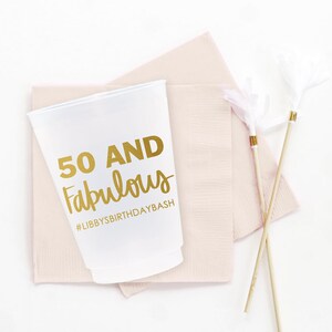 K&ouml;nnte beinhalten: Ein wei&szlig;er Plastikbecher mit goldener Aufschrift "50 and Fabulous" und "#LibbysBirthdayBash". Der Becher steht auf einer rosa Serviette.