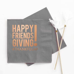 May include: Gray paper napkins with the words "HAPPY FRIENDS GIVING" in copper-colored lettering. The hashtag "#THANKFULAF" is below the text. Two decorative sticks with white feathers are also in the image.