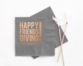 May include: Gray paper napkins with the words "HAPPY FRIENDS GIVING" in copper-colored lettering. The hashtag "#THANKFULAF" is below the text. Two decorative sticks with white feathers are also in the image.