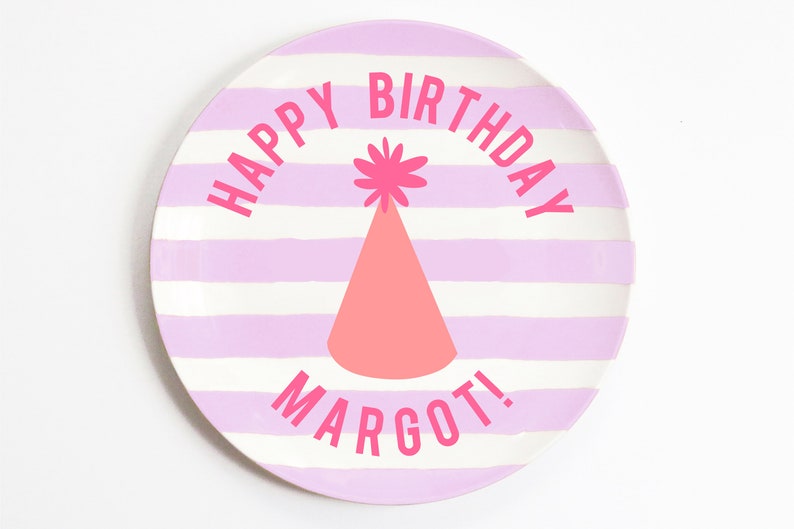 K&ouml;nnte beinhalten: Ein runder Teller mit rosa und wei&szlig;en Streifen mit dem Text "Happy Birthday Margot!" in rosa Schrift. In der Mitte des Tellers befindet sich eine rosa Partym&uuml;tze mit einem wei&szlig;en Bommel.