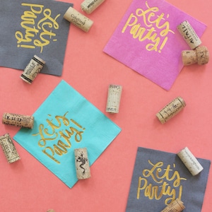Op de afbeelding: Vier grijze, blauwe en roze papieren servetten met gouden letters die "Let's Party!" zeggen. De servetten zijn omringd door kurken van wijnflessen.