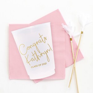 May include: White plastic cup with gold lettering that says "Congrats Kathryn! Class of 2021". The cup is sitting on a pink napkin with two white and gold feather party favors.