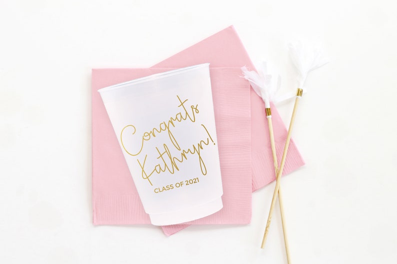 May include: White plastic cup with gold lettering that says "Congrats Kathryn! Class of 2021". The cup is sitting on a pink napkin with two white and gold feather party favors.