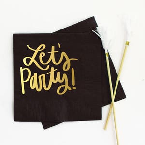Op de afbeelding: Zwarte papieren servetten met gouden letters die "Let's Party!" zeggen.
