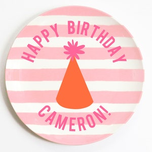 K&ouml;nnte beinhalten: Rosa-wei&szlig;er gestreifter Pappteller mit dem Text "Happy Birthday Cameron!" in Rosa. Ein rosa-oranger Partyhut mit einem rosa Bommel befindet sich in der Mitte des Tellers.