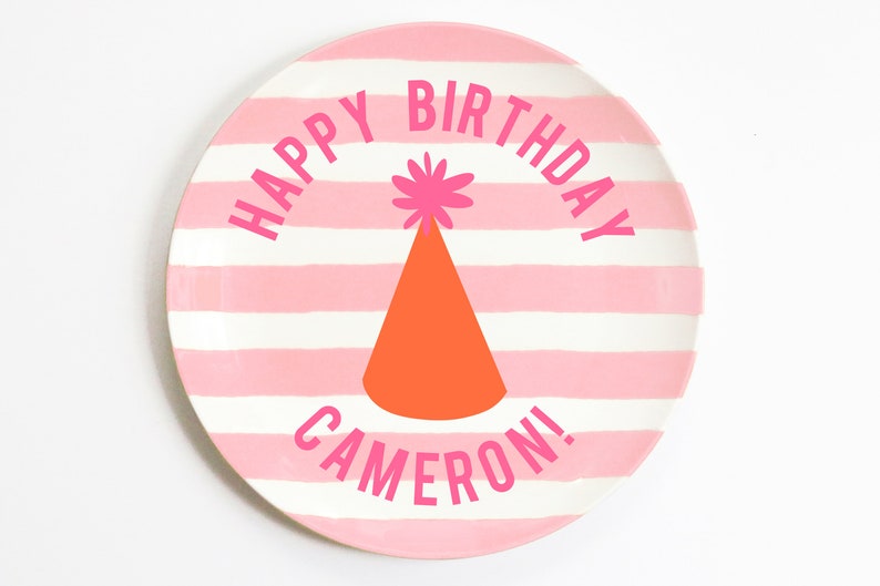 K&ouml;nnte beinhalten: Rosa-wei&szlig;er gestreifter Pappteller mit dem Text "Happy Birthday Cameron!" in Rosa. Ein rosa-oranger Partyhut mit einem rosa Bommel befindet sich in der Mitte des Tellers.
