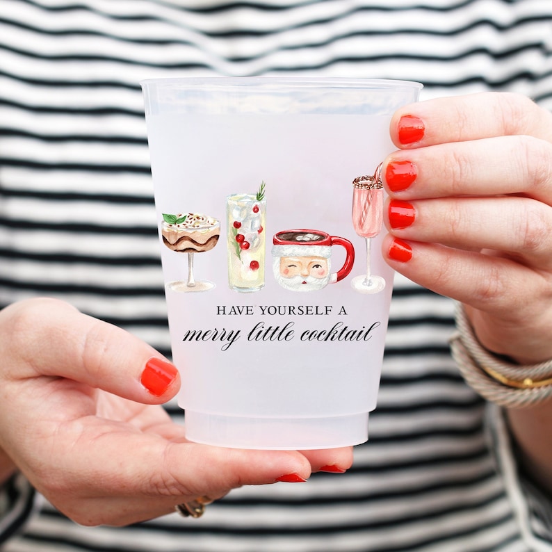 May include: A frosted plastic cup featuring watercolor illustrations of cocktails and a Santa mug. The cup has the text "Have yourself a merry little cocktail" in black script. The cup is held by a person with red nail polish.