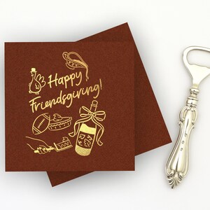 May include: Two square dark brown napkins featuring gold foil text that reads Happy Friendsgiving, accompanied by illustrations of a turkey, a football, a pie, a hand holding a serving spatula, and a wine bottle with a bow, next to a silver bottle opener.
