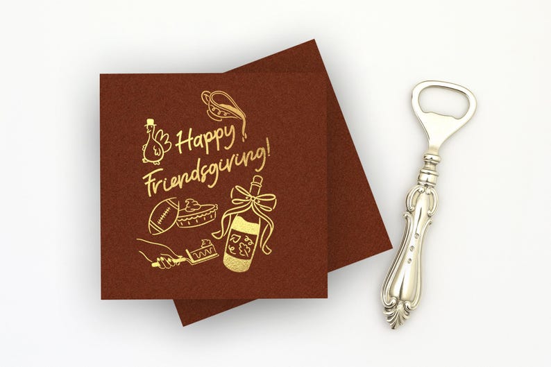May include: Two square dark brown napkins featuring gold foil text that reads Happy Friendsgiving, accompanied by illustrations of a turkey, a football, a pie, a hand holding a serving spatula, and a wine bottle with a bow, next to a silver bottle opener.