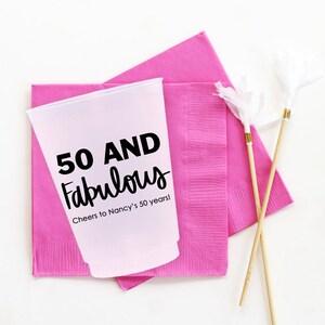 K&ouml;nnte beinhalten: Ein rosa Plastikbecher mit dem Text "50 AND Fabulous" und "Cheers to Nancy's 50 years!" darauf gedruckt. Der Becher steht auf zwei rosa Servietten. Zwei Holzspie&szlig;e mit wei&szlig;en Pompons sind ebenfalls auf dem Bild zu sehen.