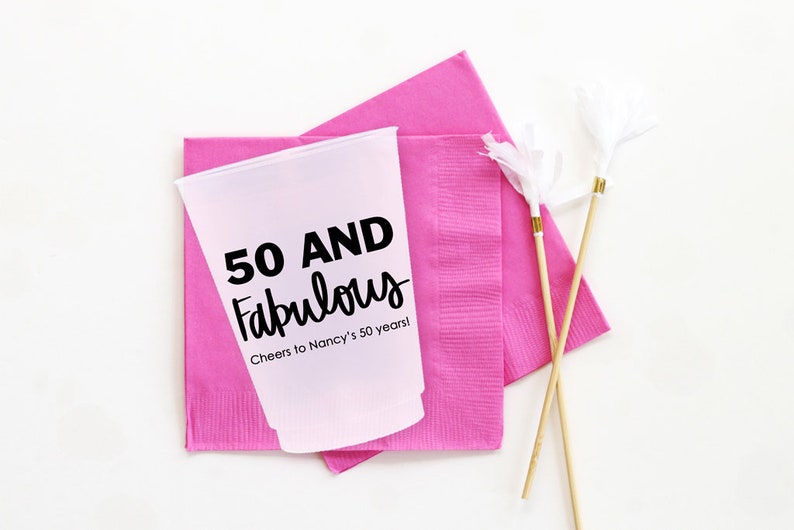 K&ouml;nnte beinhalten: Ein rosa Plastikbecher mit dem Text "50 AND Fabulous" und "Cheers to Nancy's 50 years!" darauf gedruckt. Der Becher steht auf zwei rosa Servietten. Zwei Holzspie&szlig;e mit wei&szlig;en Pompons sind ebenfalls auf dem Bild zu sehen.