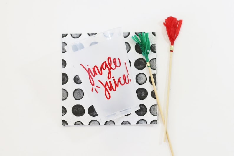 May include: A clear plastic cup with the words "Jingle Juice!" in red lettering sits on a black and white polka dot napkin. Two decorative wands with red and green tassels are also present, suggesting a festive theme.