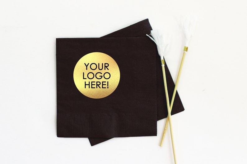 May include: Two black paper napkins with a gold circle sticker that says "YOUR LOGO HERE!" on top of each other. The napkins are on a white surface with two wooden skewers with white fluffy pom-poms on the ends.