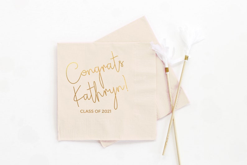 May include: Pink paper napkins with gold foil lettering that reads "Congrats Kathryn! Class of 2021".