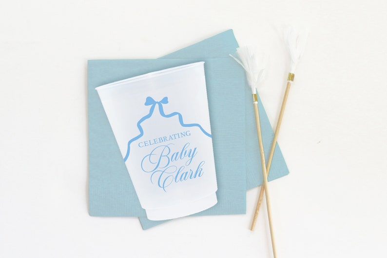 May include: A white paper cup with a blue ribbon design and the text "Celebrating Baby Clark". The cup is on top of a light blue napkin. Two white feather-tipped sticks are on a light blue napkin.