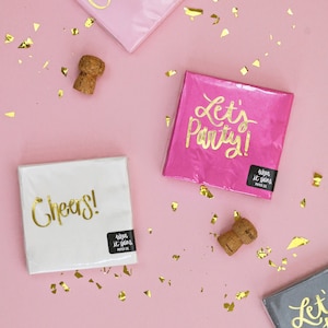 Op de afbeelding: Vier roze en witte papieren servetten met gouden letters. De servetten zeggen "Cheers!" en "Let's Party!" De servetten liggen op een roze achtergrond met gouden confetti.