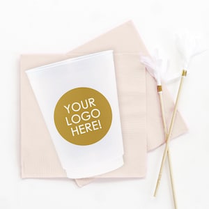 May include: A white plastic cup with a gold circle sticker that says "YOUR LOGO HERE!" on a pink napkin. Two white feather cocktail stirrers are on the right side of the image.