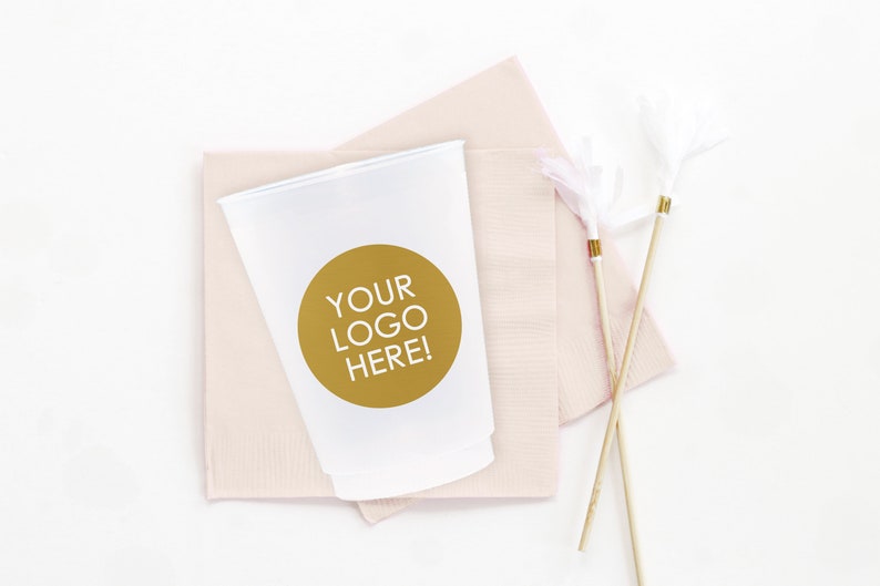 May include: A white plastic cup with a gold circle sticker that says "YOUR LOGO HERE!" on a pink napkin. Two white feather cocktail stirrers are on the right side of the image.